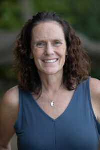 A smiling woman with curly hair, wearing a blue sleeveless top and a necklace, poses outdoors.