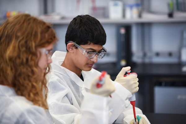 Two students in lab coats and goggles are engaged in a science experiment, using pipettes in a laboratory setting.