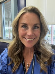 A smiling woman with long, wavy hair, wearing a blue top and a necklace, poses in a well-lit room.