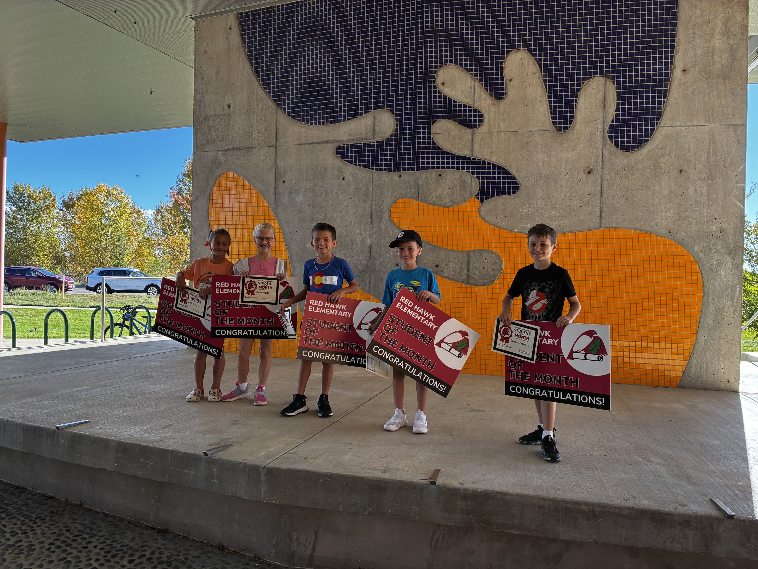 Five children stand on a stage holding "Student of the Month" signs, with colorful wall art and trees in the background.